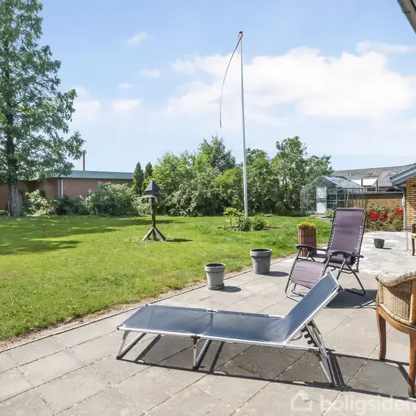 Solstole står på en terrasse af fliser, omkranset af en velplejet græsplæne og frodige buske. I baggrunden ses et flagstang og et fuglehus.