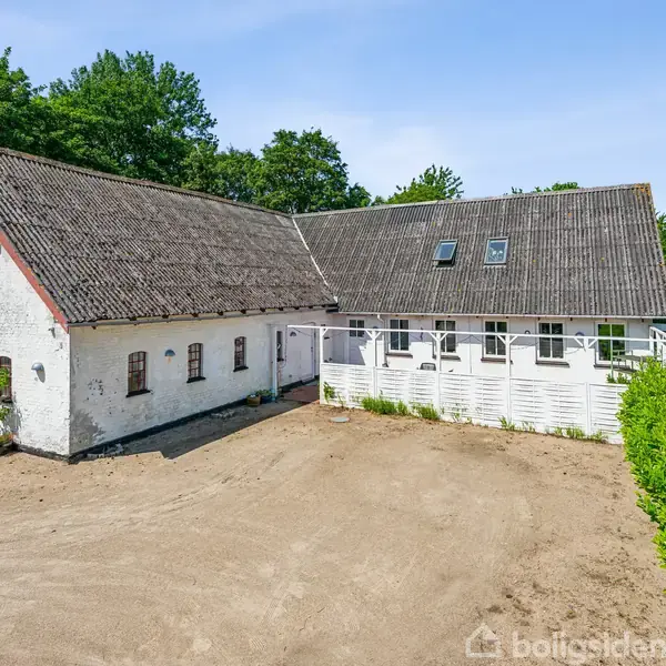 Hvid landhusbygning med stråtækt tag, forbundet med en nyere udvidelse til højre. Omkring bygningen er der en grusbelagt gårdsplads og frodig grøn vegetation i baggrunden.