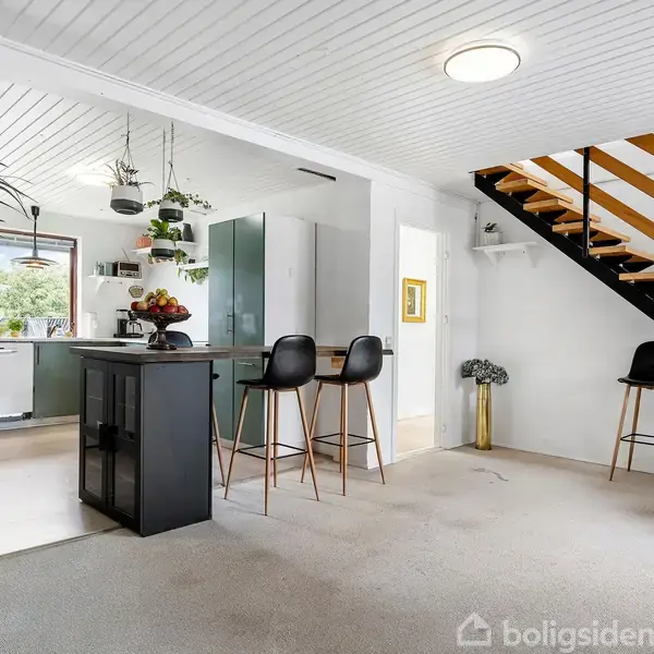 Et køkken-alrum med en køkkenø og tre barstole. En åben trappe fører til en anden etage. Området er lyst og moderne, med planter og dekorative elementer.