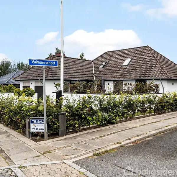 Hus til salg. Skiltet viser "Valmuemvænget." Huset har et mørkt tegltag og hvide vægge, omgivet af hæk. Placeret på et hjørne af en stille vej. Telefonnummer: 76 804 804.