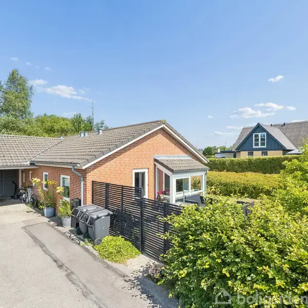 Et murstenshus med gråt tag står ved siden af en parkeringsplads og indkørsel. Omgivet af hække og træer i et boligområde med klart, blåt himmel i baggrunden.