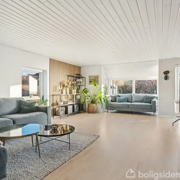 Stue med grå sofaer og sofabord, dekoreret med planter i krukker og kunst på væggene. Rummet har store vinduer, der giver indgang til naturligt lys og udsigt til haven.