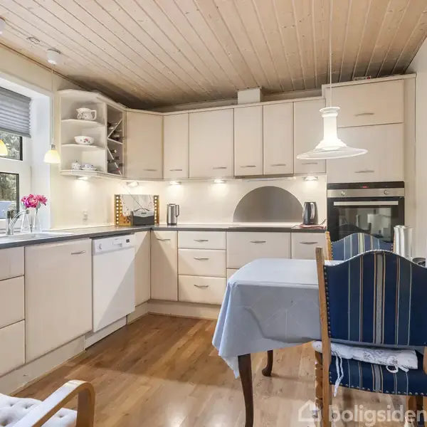 Køkken med lyse skabe og spisebord dækket med blåt stof. Køkkenmaskiner står på bordpladen, og en lampe hænger ned fra loftet. Vindue med persienne og udsigt til træer.