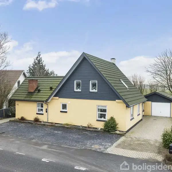 Et gult hus med sort gavl står ved siden af en vej. Det har et grønt tag og to ovenlysvinduer. Der er en garage til højre og åbne marker i baggrunden.