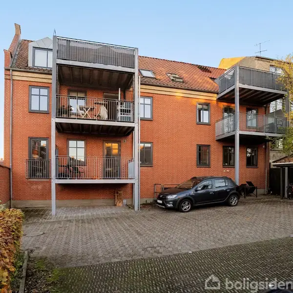 En rød murstensbygning med tre altaner i flere etager. På parkeringspladsen står en bil og nogle cykler. Omgivelserne indeholder en brostensbelægning og beplantning med efterårsfarver.