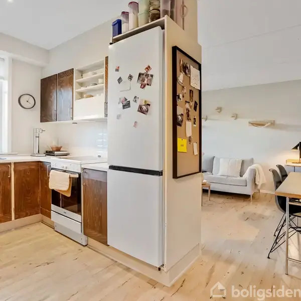 Et køkken med køleskab fyldt med magneter og billeder, ved siden af en spiseplads med et bord og stole. Stuen i baggrunden har en sofa og dekorationer.