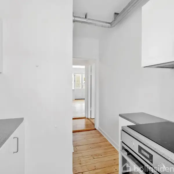 Et køkken med sorte bordplader og hvide skabe, set fra en vinkel, der viser en gang fører til et lyst rum med trægulv i baggrunden.