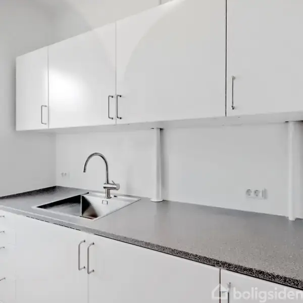 Hvide køkkenskabe hænger på en væg over en grå bordplade med en indbygget stålvask og vandhane. Sollys strømmer ind gennem et vindue til venstre.