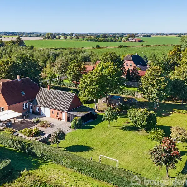 Et rødt murstenshus med en terrasse ses fra oven. Omkring huset er en stor, velplejet have med træer og hække. Landskabet omkring består af marker og skove i det fjerne.