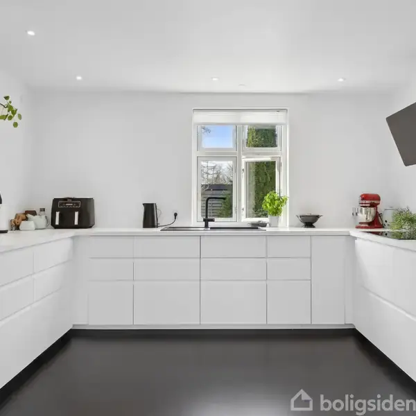 Et moderne køkken med hvide skabe og sorte apparater. En stor, lysende vindue i midten omkranset af planter og køkkenudstyr skaber en stilfuld, ren atmosfære.