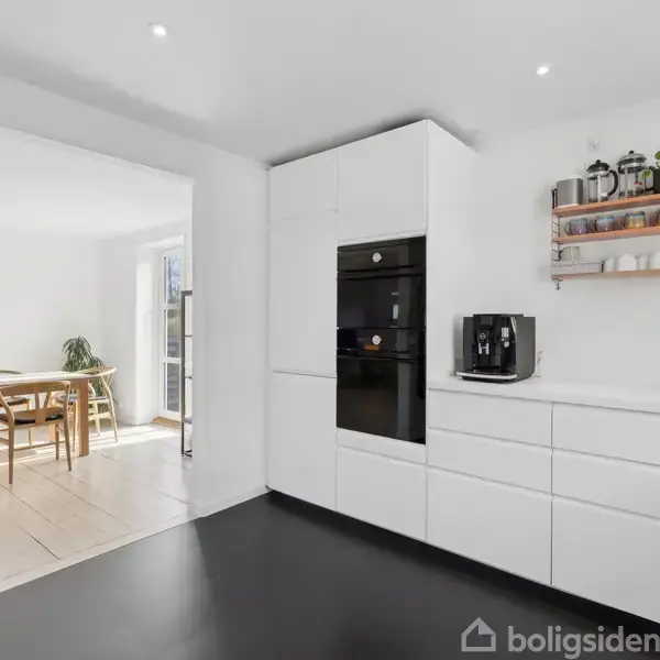 Hvidt, moderne køkken med indbygget ovn og skuffer. Kaffemaskine på bordpladen og hylde med planter og krukker ovenfor. I baggrunden ses en lys spisestue med træmøbler.