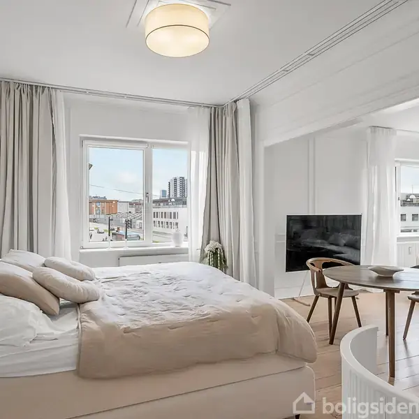 En seng står i et lyst værelse med en rund spisebord og stole ved siden af. Store vinduer giver udsigt over byens skyline, med gardiner langs siderne.