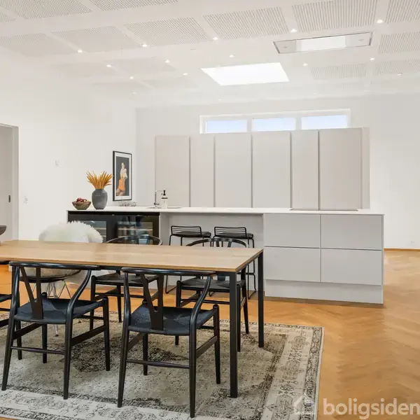 Et moderne køkken med en stor, lys bordplade og tre barstole foran. Et spisebord med seks stole står på et mønstret tæppe på trægulv. Hvide vægge og loft.
