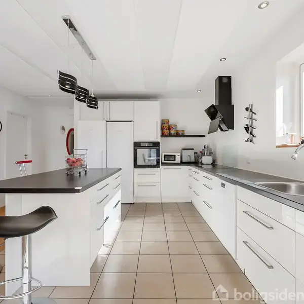 Et moderne køkken med hvide skabe og sorte bordplader. Midt i rummet er en køkkenø med barstole. Der er store vinduer, masser af lysindfald og et vægur på hvid væg.