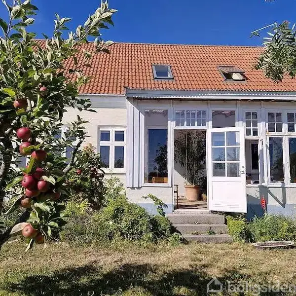 Et hus med rødt tegltag og store vinduer står i en baghave. Dør er åben. Foran er et æbletræ med røde æbler. Græsset er grønt.
