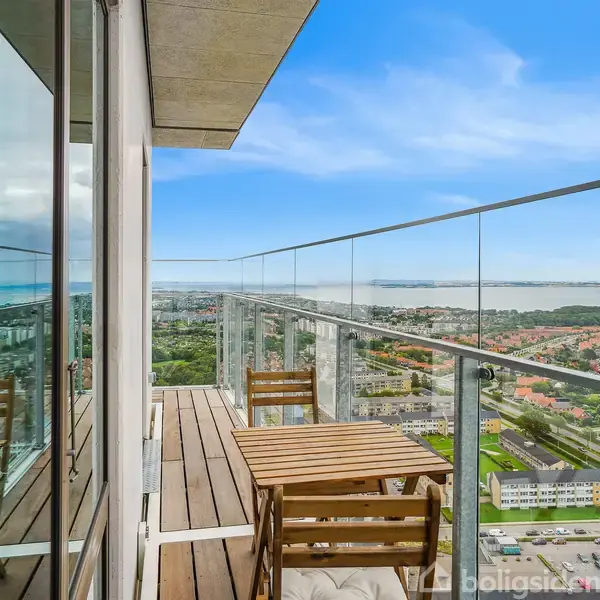 Et bord og to stole står på en balkon med glasrækværk, reflektérende i vinduerne. De har udsigt over en by med kystlinje i baggrunden under en blå himmel.