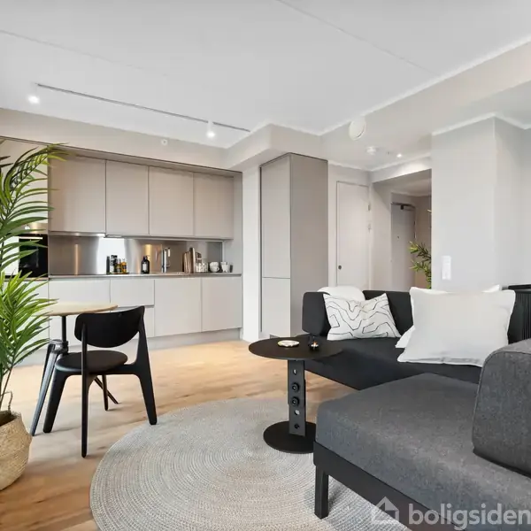 En grå sofa står i et moderne åbent køkken/alrum med trægulv. En sort stol og bord står op ad en beige væg med indbygget køkken. Grøn plante ved vindue.