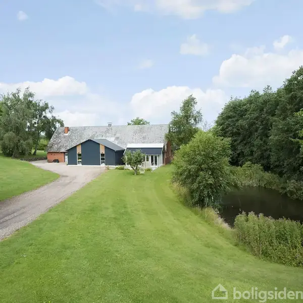 Et landsted ligger ved en grusvej omgivet af grønne træer og græsarealer. Til højre ses en dam, og himlen er delvist skyet med blå nuancer.