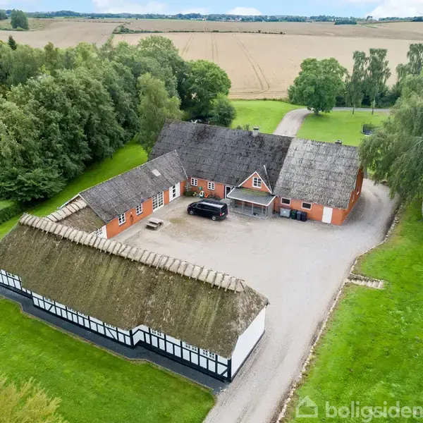 Et stråtækt bondehus med en gårdsplads og en bil parkeret foran. Omkranset af grønne marker og træer, med en grusvej der fører til bygningen.
