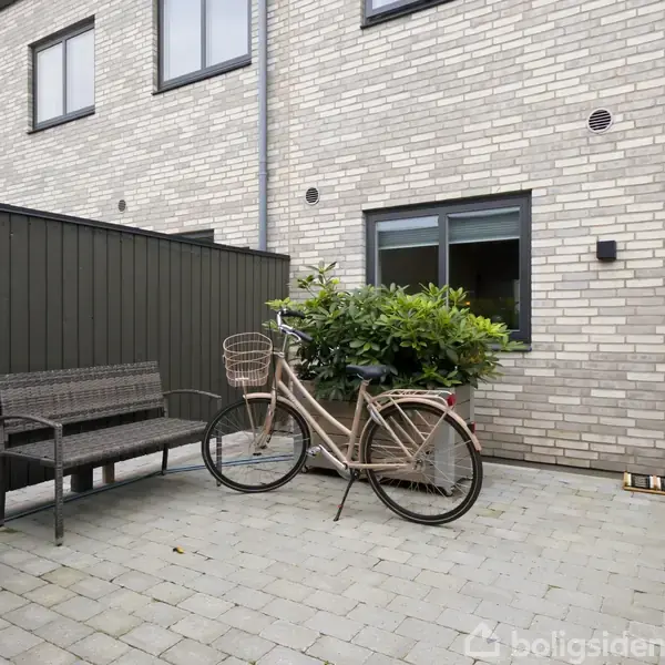 Cykel med kurve parkeret ved en murstensbygning, omkranset af en plante i baggrunden. Ved siden af står en bænk og en plantekrukke på en flisebelagt gårdsplads.