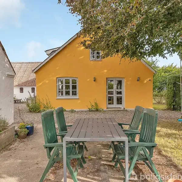 Et bord med seks grønne stole står på en terrasse. Foran ses et gult hus med en havedør og vinduer. Træer omkranser gårdhaven.