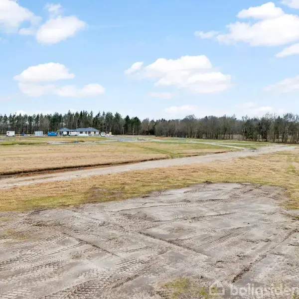 En åben mark med tør jord og grus ligger forrest. En vej løber gennem området, ledsaget af få spredte træer og buske. Himlen er klar med spredte skyer.