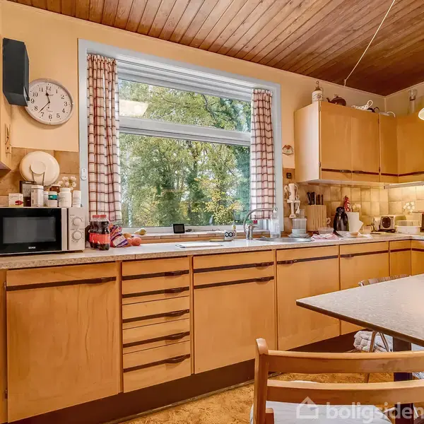 Køkken med beige træskabe, mikroovn og komfur. Bord med stoler ved siden. Vindue med ternet gardin viser træer udenfor. Vægmæssig ure i hjørnet. Ingen tekst fundet.