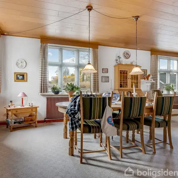 Et spisebord med seks stole står midt i en lys stue. Bordet er dækket med en dug, og over det hænger to lamper. Møbler inkluderer kommoder og bogreoler.