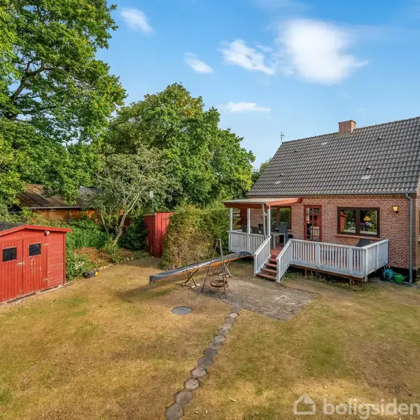 En rød murstenshus med en terrasse ligger i en have med græsplæne og træer. Ved siden af huset ses et rødt skur.