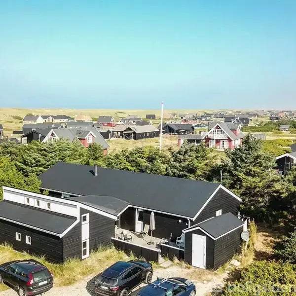 Sort hus med terræn i skovområde omringet af andre huse, nær kysten. Flere biler parkeret foran. Klar blå himmel dominerer landskabet med havet i horisonten.
