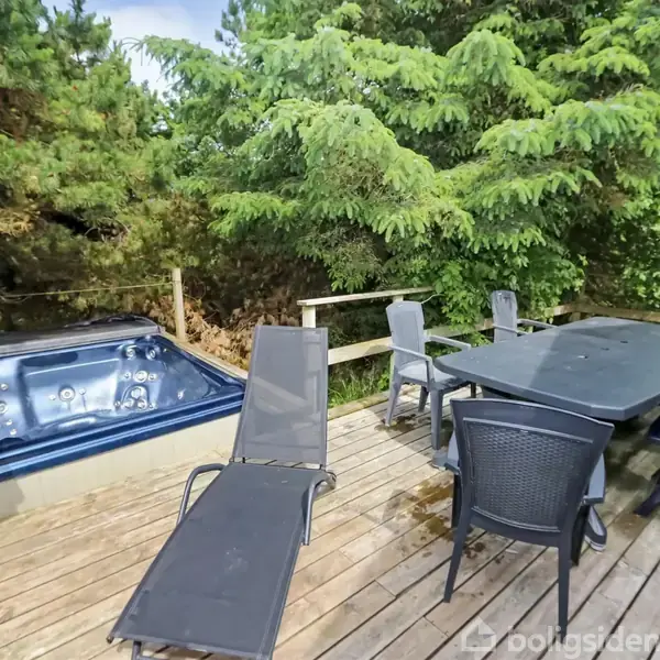 Jacuzzi står ubrugt på en træterrasse omgivet af grønne træer. Ved siden af er der en udendørs spiseplads med sort bord og stole, skaber en afslappet atmosfære.