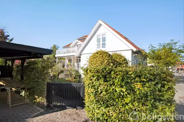 Et hvidt hus med rødt tegltag står i en solrig have omgivet af grønne buske. Til venstre ses en sort træcarport. Himlen er klar og blå.