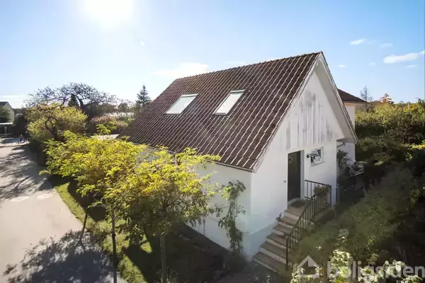 Et hvidt hus med tegltag har en trappe, der fører op til hoveddøren. Omgivet af frodige træer og buske i et solrigt boligområde.