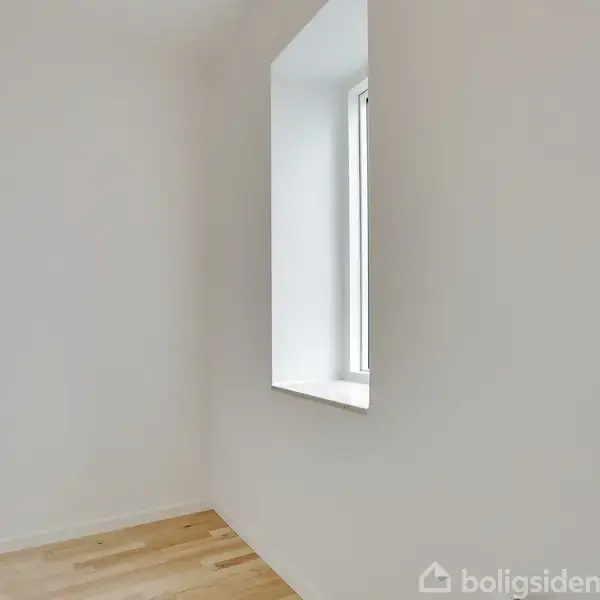 Et minimalistisk rum har et stort vindue, der lader naturligt lys slippe ind. Rummets vægge er hvide, og gulvet er træ, hvilket skaber en lys og enkel atmosfære.