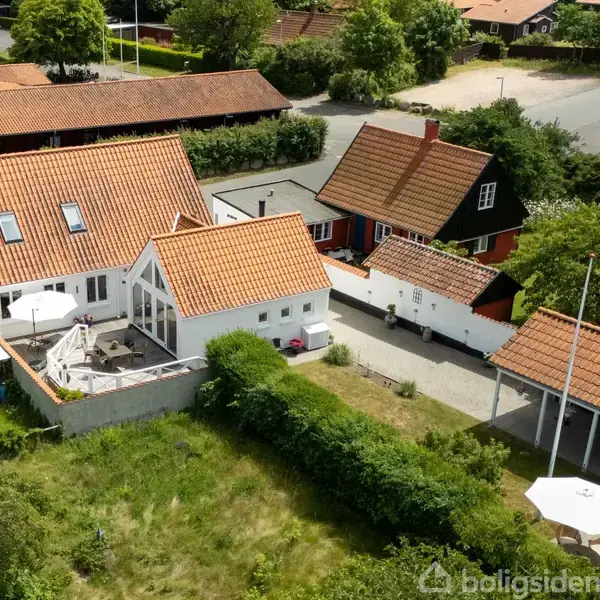 Hus med rødt tegltag og terrasse ses ovenfra i et villakvarter. Haven er omgivet af buske og træer. Rundt om huset er der flere mindre bygninger og en grusvej.