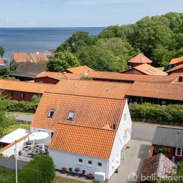 Et hvidt hus med rødt tegltag står i fokus. Det omkranses af flere huse og grønne træer, med havet i baggrunden. En veranda er synlig.