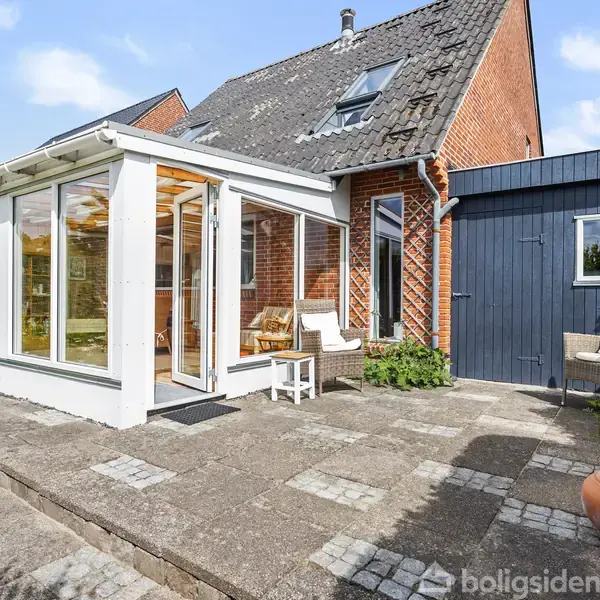 En moderne udestue med store vinduespartier er bygget til et rødt murstenshus. Terrassen foran udestuen har møbler og planter. Blåt skur ses ved siden af huset i solrigt vejr.