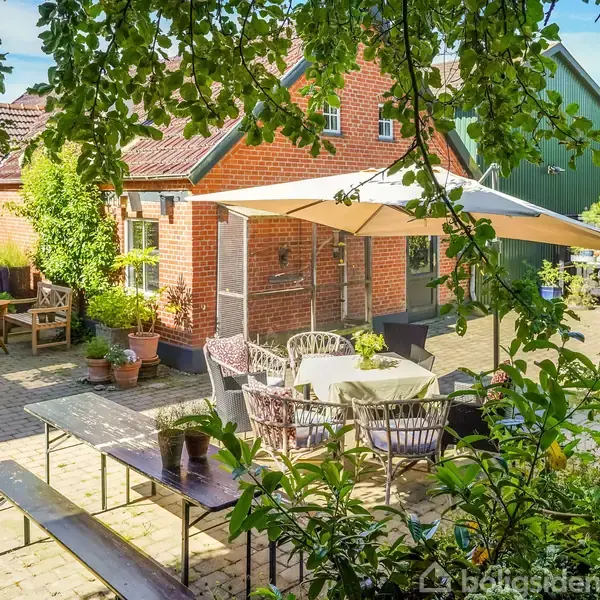 Terrassemøbler står under en stor parasol på en stenbelagt gårdsplads foran et rødt murstenshus. Omkring møblerne er der planter i krukker og ekstra siddepladser.