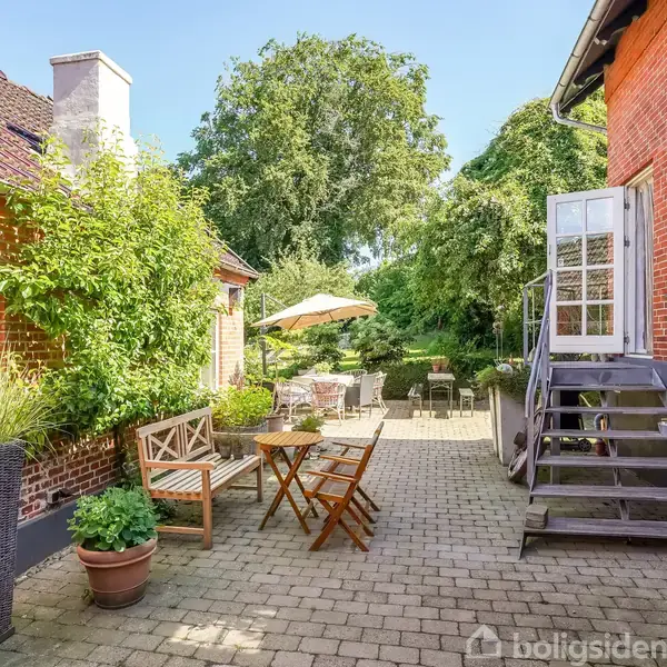 Terrasse med havemøbler og grønne planter, omgivet af murstensbygninger. Trappe fører op til en dør. Parasol og bordarrangement står i baggrunden under et stort træ.