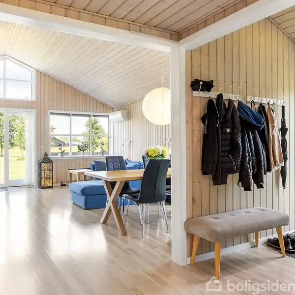 Entré med knagerække fyldt med jakker og en bænk ved siden af hoveddør. I baggrunden ses en stue med spisebord, blå sofa, hvide vægge og store vinduer med udsigt til have.