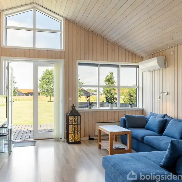 Stue med blå hjørnesofa står i et lyst rum med store vinduer og loft til kip. Udenfor ses en grøn plæne og træer gennem en glasdør. En lanterne står ved vinduet.