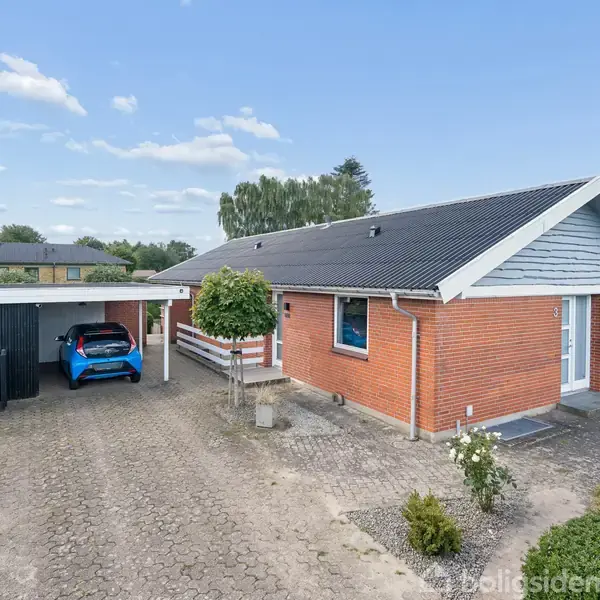 Rødt murstenshus med gråt sadeltag, stående stille, omgivet af en have. En blå bil holder parkeret i carporten. Indkørsel med fliser fører til husets hovedindgang.