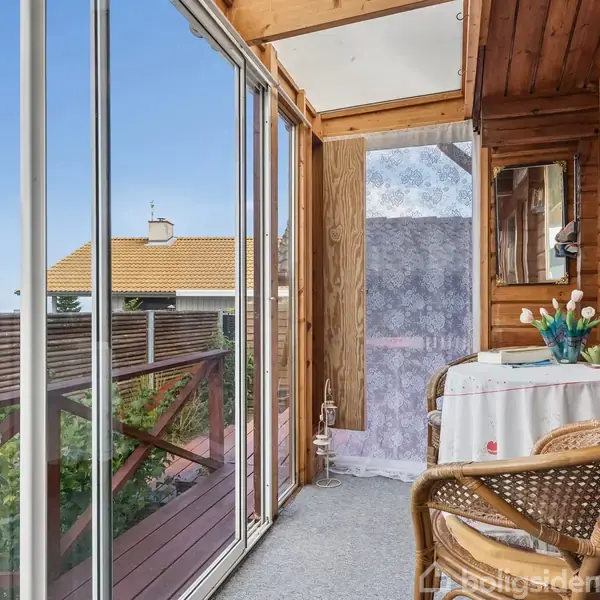 En overdækket balkon med trævægge og store glaspartier viser en udsigt til et hus med gult tag. Et lille bord med dug og træstol står i hjørnet. Blomsterdekoration pryder bordet.
