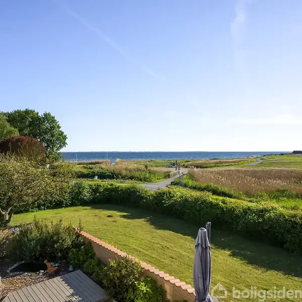 En have med terrasse og græsplæne strækker sig ud mod en sti, der fører til havet. Landskabet er grønt med træer og buske. Sæsonen ser ud til at være sommer.