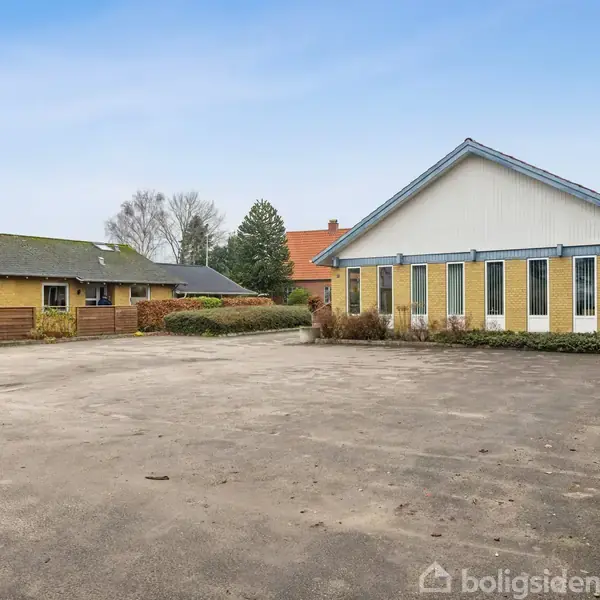 En gul bygning står tom på en stor asfalteret plads, omgivet af buske og træer under en blå himmel. En anden gul bygning ses til venstre.