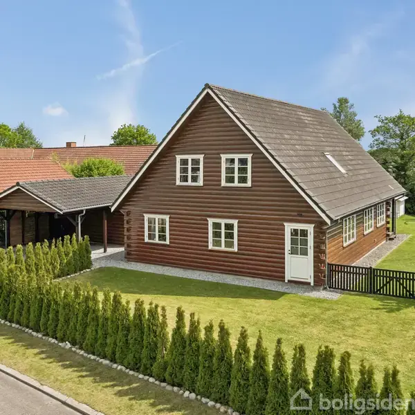 Brun træhus med hvide vinduer står stille i et velholdt haveområde. Foran ses en velplejet hæk og en grusindkørsel. Naboen har et rødt tag, og en flagstang står i baggrunden.