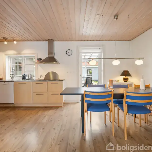 Et moderne køkken med lyse træskabe og indbygget komfur. Ved siden af står et spisebord med blå stole, der ligger i et rum med trægulv og hvide vægge. Ingen tekst.