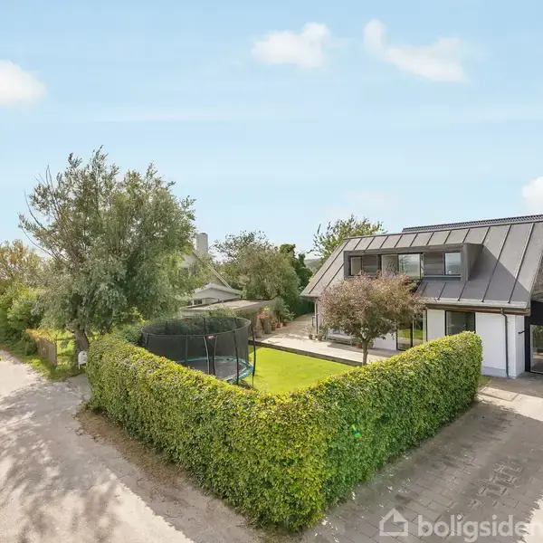 Hus med moderne arkitektur omgivet af en velplejet have og hække; trampolin placeret på græsplænen. Naboer ses langs den stille vej i et boligområde med træer.