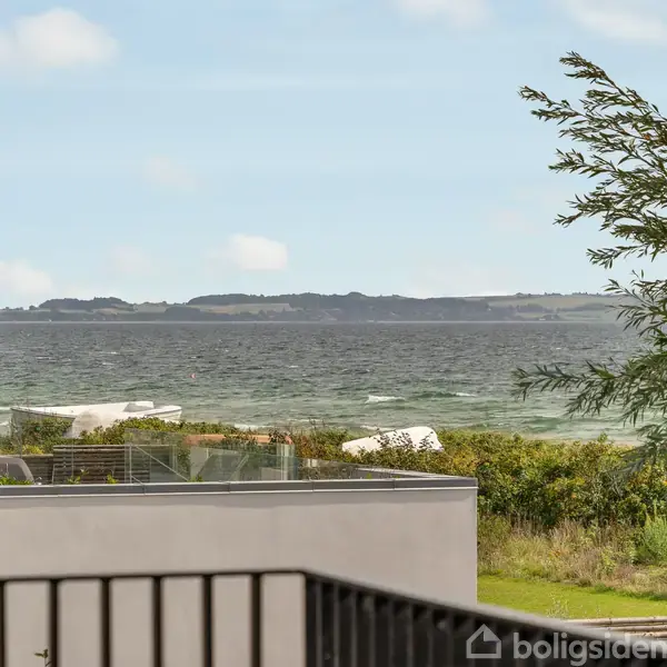 Udsigt over havet fra en balkon med sort gelænder; huse og buskadser forekommer i forgrunden. Baggrunden viser en horisont med skyer over vandet.