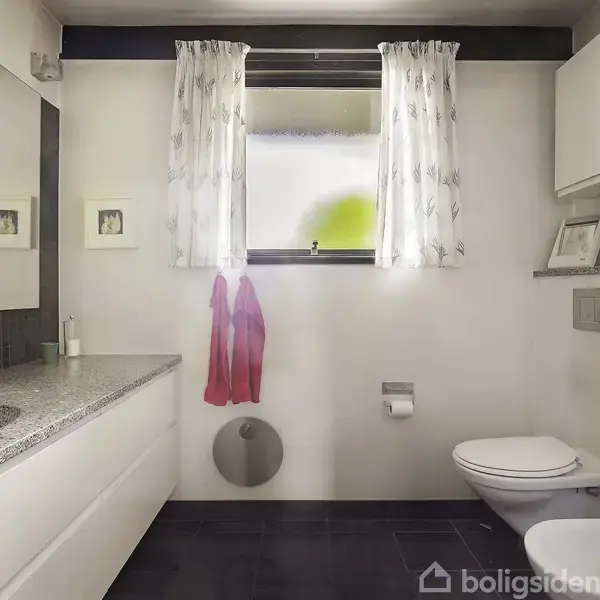 Badeværelse med hvidt møblement og sort gulv. Spejl over stor håndvask. Toilet til højre under et vindue med gardiner. Toiletrulleholder og håndklæder ved siden af.
